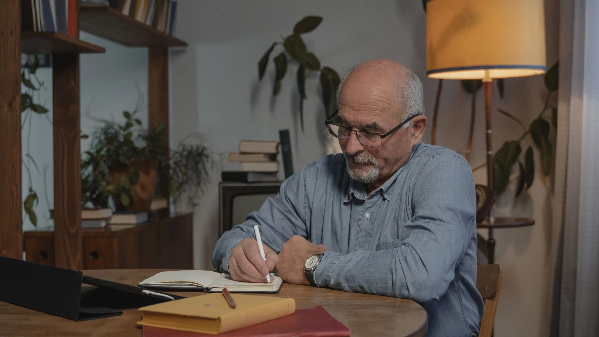 An older man writes notes in a notebook at a table in a cozy living room