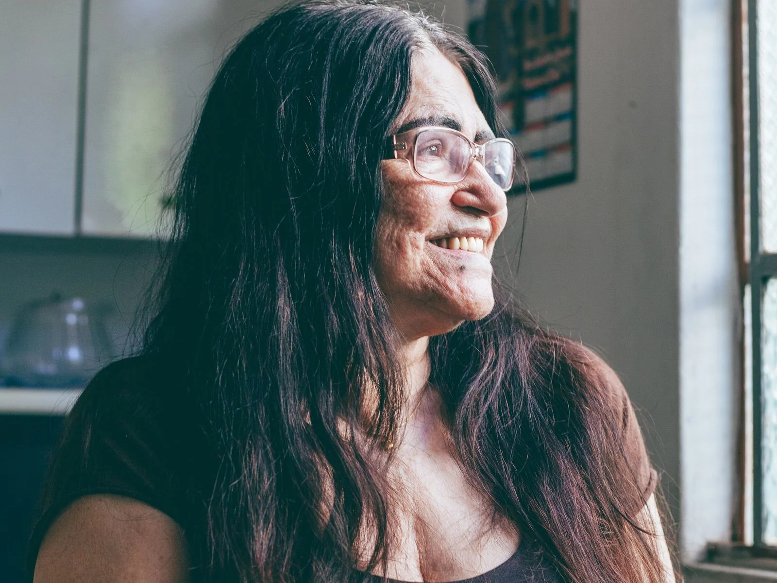 An older woman with glasses and long dark hair smiles while sitting near a bright window at home.